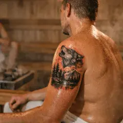 Mann mit Wolf-Tattoo in der Sauna. Dampf steigt von den heißen Steinen auf.