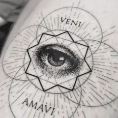 Ein filigranes Fineline Tattoo am Oberarm einer Frau zeigt ein Auge inmitten geometrischer Formen und der Worte "VENI AMAVI".