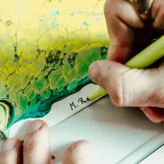 Künstler setzt seine Unterschrift unter ein abstraktes Gemälde in Gelb- und Grüntönen.