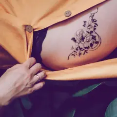 Ein zartes Tatoo mit einer Blume ziert die Haut, umrahmt von einem gelben Stoff und grünen Blättern.