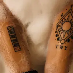 Zwei Arme mit Tattoos in Schaum gebadet. Ein Tattoo zeigt ein geometrisches Muster mit Sonne und Mond, das andere eine Reihe von Symbolen. PH neutrale Seife Tattoo-Pflege.