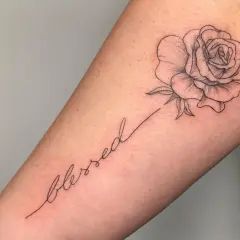 Ein zarter Fineline-Schriftzug "blessed" mit einer kleinen Rose als Tattoo.