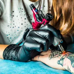 Künstler mit schwarzen Handschuhen arbeitet an einem Tattoo auf dem Arm. Ein rotes Tattoo-Gerät wird verwendet, um ein neues Design zu erstellen.