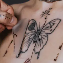 Ein Mann mit tätowierter Hand zieht eine Schutzfolie von einem frisch gestochenen Schmetterling-Tattoo auf der Brust ab. Pflege danach ist wichtig.