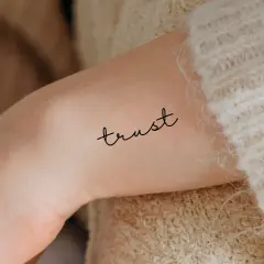 Ein zartes Tattoo mit dem Wort "trust" (Vertrauen) auf einem Arm, umgeben von weichem Strick.