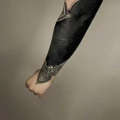 Ein Arm mit einem beeindruckenden **blackout tattoo**, das fast vollständig in tiefem Schwarz gehalten ist. Zarte Mandalas schmücken Handgelenk und Ellenbogen.