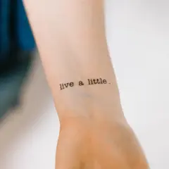 Ein Tattoo mit der Aufschrift "live a little" in Schreibmaschinenschrift auf einem Handgelenk.