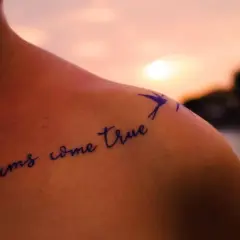 Schulter einer starken Frau mit einem Spruch-Tattoo: "dreams come true". Ein Vogel fliegt über den Schriftzug.