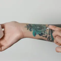 Pflege für dein Tattoo: Eine Hand hält eine Dose mit weißer Creme, während die andere Hand sie auf ein buntes Tattoo aufträgt. Kokosöl Tattoo-Pflege.