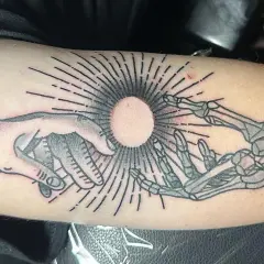Ein Tattoo zeigt eine menschliche Hand und eine Skeletthand, die sich auf einen hellen Kreis zubewegen. Die Haut ist leicht gerötet, als ob sie gerade erst aufgerissen wurde, um das Kunstwerk zu enthüllen.