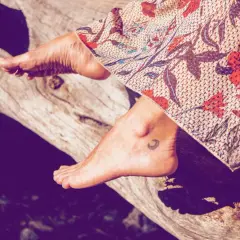 Ein Fuß mit einem kleinen Mond-Tattoo ruht auf einem Baumstamm, bedeckt von einem gemusterten Stoff.