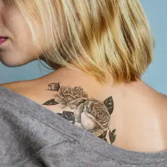 Ein frisches Tattoo mit Rosen auf der Schulter einer Frau, das mit einem grauen Pullover abgedeckt wird.
