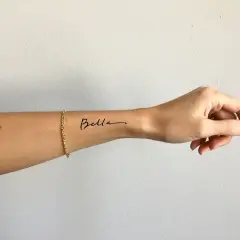 Ein zartes Tattoo-Armband mit dem Namen "Bella" auf dem Handgelenk, geschmückt mit einer goldenen Kette.