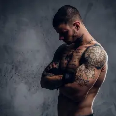 Ein muskulöser Mann mit vielen Tattoos, der seine Arme verschränkt. Sein Körperbau zeigt, dass er viel für Sport und Training tut.
