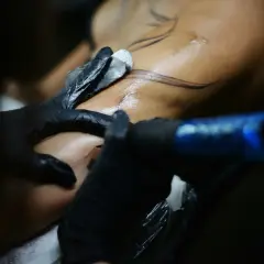 Detailaufnahme: Tätowierer mit schwarzen Handschuhen arbeitet an einem Arm. Feine Linien für blackwork tattoos werden auf die Haut gebracht.