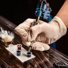 Ein Mann bekommt ein Knie Tattoo. Die Tätowiermaschine taucht in schwarze Tinte.