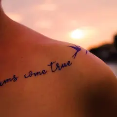 Schulter mit Tattoo "dreams come true" in geschwungenen Tattoo Schriftarten, daneben eine kleine Schwalbe.