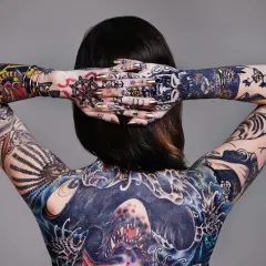 Rückenansicht einer Frau mit aufwendigen, abstrakten Tattoos, die ihren ganzen Körper bedecken. Goldene Nägel und Schmuck ergänzen die Kunstwerke.