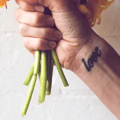Ein Handgelenk mit einem "Love"-Tattoo, das an ein Freundschaftstattoo erinnert, hält einen Strauß gelber Blumen.