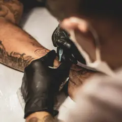 Tätowierer mit schwarzen Handschuhen arbeitet an einem Arm. Ein detailliertes Tattoo ohne Schattierung entsteht.