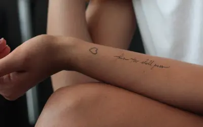 Ein Tattoo auf dem Unterarm einer Frau mit der Aufschrift "this too shall pass" und einem kleinen Herz. Ein Symbol der Hoffnung nach der Überwindung von Alkoholsucht.