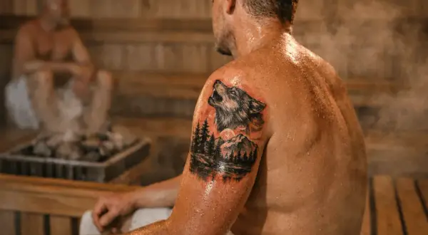 Mann mit Wolf-Tattoo in der Sauna. Dampf steigt von den heißen Steinen auf.
