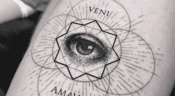 Ein filigranes Fineline Tattoo am Oberarm einer Frau zeigt ein Auge inmitten geometrischer Formen und der Worte "VENI AMAVI".