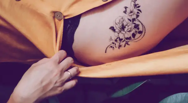 Ein zartes Tatoo mit einer Blume ziert die Haut, umrahmt von einem gelben Stoff und grünen Blättern.