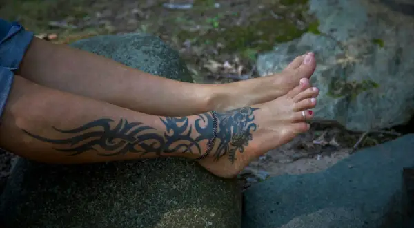 Beine mit einem Tribal-Tattoo, das sich über Wade und Fuß zieht. Das Tattoo kribbelt, als die Person auf einem Stein sitzt.