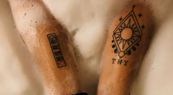Zwei Arme mit Tattoos in Schaum gebadet. Ein Tattoo zeigt ein geometrisches Muster mit Sonne und Mond, das andere eine Reihe von Symbolen. PH neutrale Seife Tattoo-Pflege.