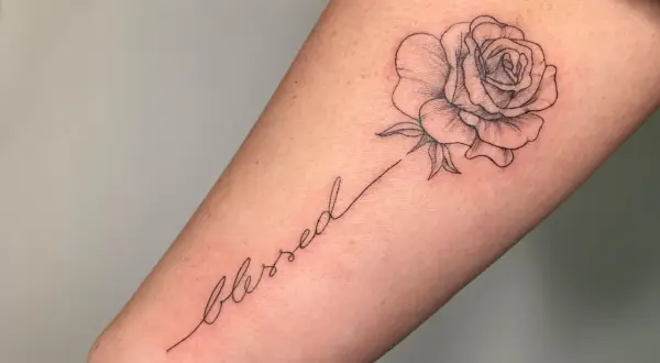Ein zarter Fineline-Schriftzug "blessed" mit einer kleinen Rose als Tattoo.