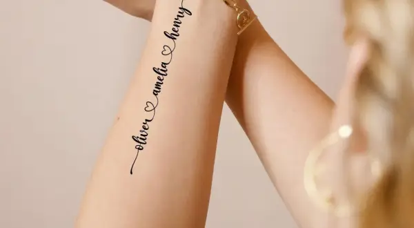 Ein zartes **Enkel Tattoo Namen** zeigt "oliver & amelia & henry" in geschwungener Schrift auf einem Arm, geschmückt mit einem goldenen Armband.