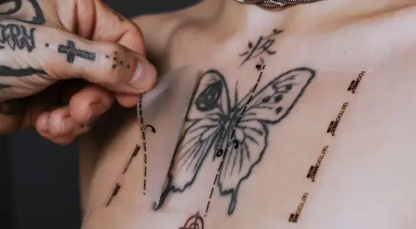 Ein Mann mit tätowierter Hand zieht eine Schutzfolie von einem frisch gestochenen Schmetterling-Tattoo auf der Brust ab. Pflege danach ist wichtig.
