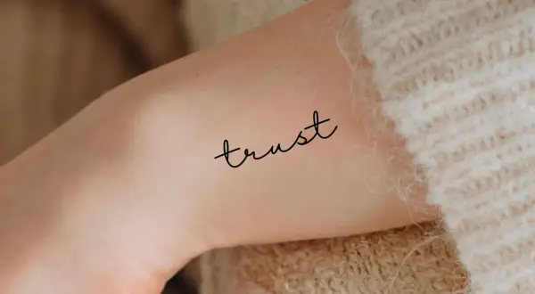 Ein zartes Tattoo mit dem Wort "trust" (Vertrauen) auf einem Arm, umgeben von weichem Strick.