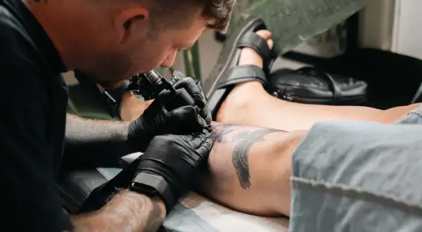 Tätowierer arbeitet an einem Adler-Motiv auf dem Schienbein einer Frau. Tolle Ideen für ein Tattoo am Schienbein einer Frau.
