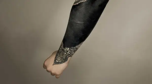 Ein Arm mit einem beeindruckenden **blackout tattoo**, das fast vollständig in tiefem Schwarz gehalten ist. Zarte Mandalas schmücken Handgelenk und Ellenbogen.