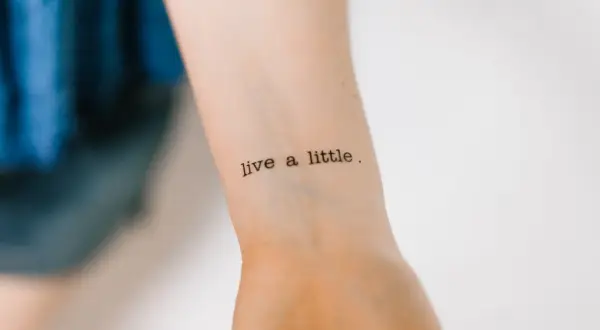 Ein Tattoo mit der Aufschrift "live a little" in Schreibmaschinenschrift auf einem Handgelenk.