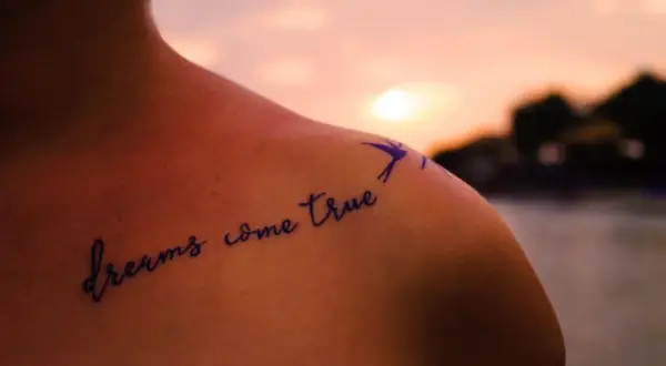 Schulter einer starken Frau mit einem Spruch-Tattoo: "dreams come true". Ein Vogel fliegt über den Schriftzug.