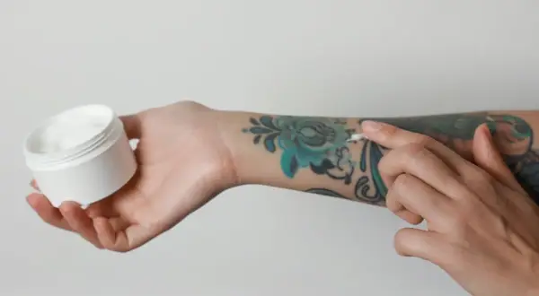 Pflege für dein Tattoo: Eine Hand hält eine Dose mit weißer Creme, während die andere Hand sie auf ein buntes Tattoo aufträgt. Kokosöl Tattoo-Pflege.