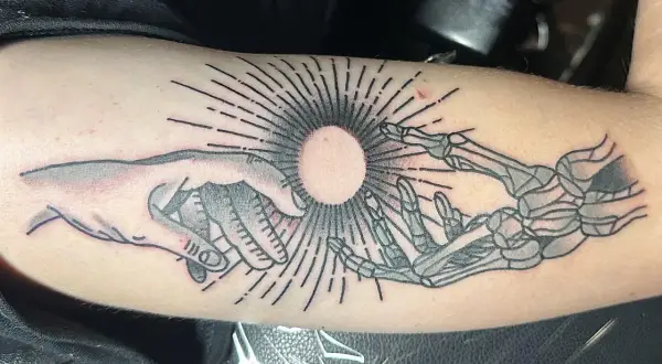 Ein Tattoo zeigt eine menschliche Hand und eine Skeletthand, die sich auf einen hellen Kreis zubewegen. Die Haut ist leicht gerötet, als ob sie gerade erst aufgerissen wurde, um das Kunstwerk zu enthüllen.