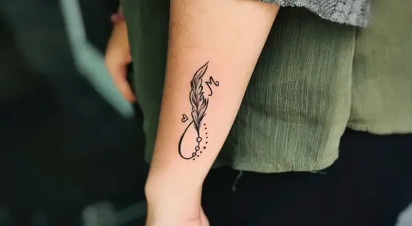 Ein kleines Feder-Tattoo mit Unendlichkeitszeichen und dem Buchstaben M auf dem Unterarm eines Mannes.