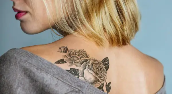 Ein frisches Tattoo mit Rosen auf der Schulter einer Frau, das mit einem grauen Pullover abgedeckt wird.