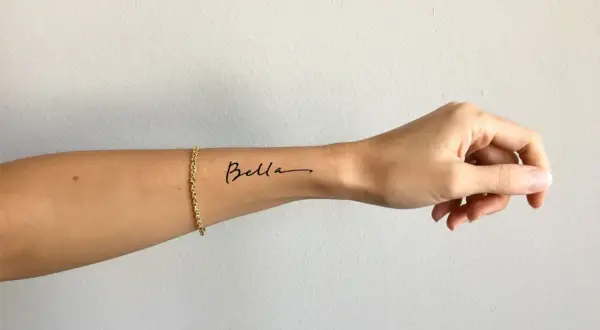 Ein zartes Tattoo-Armband mit dem Namen "Bella" auf dem Handgelenk, geschmückt mit einer goldenen Kette.