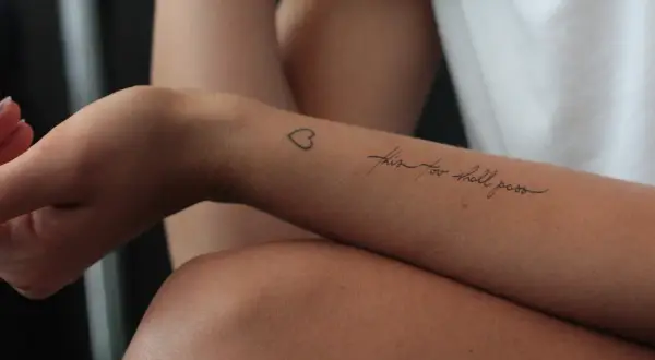 Ein Tattoo auf dem Unterarm einer Frau mit der Aufschrift "this too shall pass" und einem kleinen Herz. Ein Symbol der Hoffnung nach der Überwindung von Alkoholsucht.