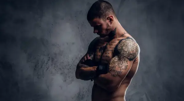 Ein muskulöser Mann mit vielen Tattoos, der seine Arme verschränkt. Sein Körperbau zeigt, dass er viel für Sport und Training tut.