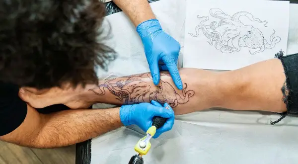 Tätowierer mit blauen Handschuhen sticht Oktopus-Motiv auf Bein. Tolle Tattoo-Ideen für Meeresliebhaber.