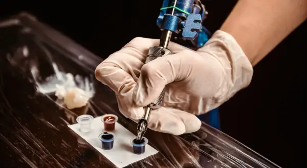 Ein Mann bekommt ein Knie Tattoo. Die Tätowiermaschine taucht in schwarze Tinte.