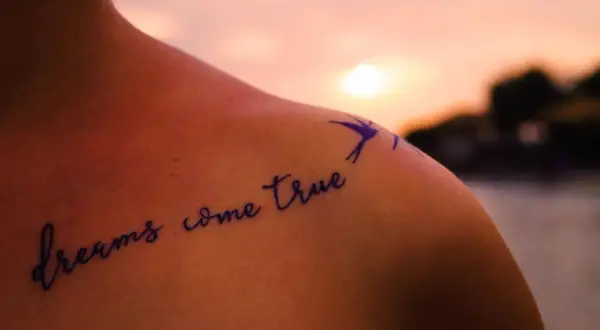 Schulter mit Tattoo "dreams come true" in geschwungenen Tattoo Schriftarten, daneben eine kleine Schwalbe.