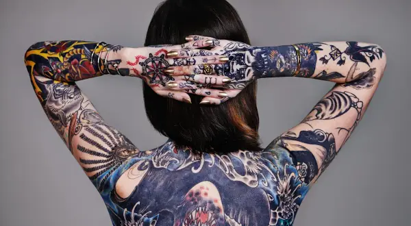Rückenansicht einer Frau mit aufwendigen, abstrakten Tattoos, die ihren ganzen Körper bedecken. Goldene Nägel und Schmuck ergänzen die Kunstwerke.