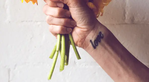 Ein Handgelenk mit einem "Love"-Tattoo, das an ein Freundschaftstattoo erinnert, hält einen Strauß gelber Blumen.